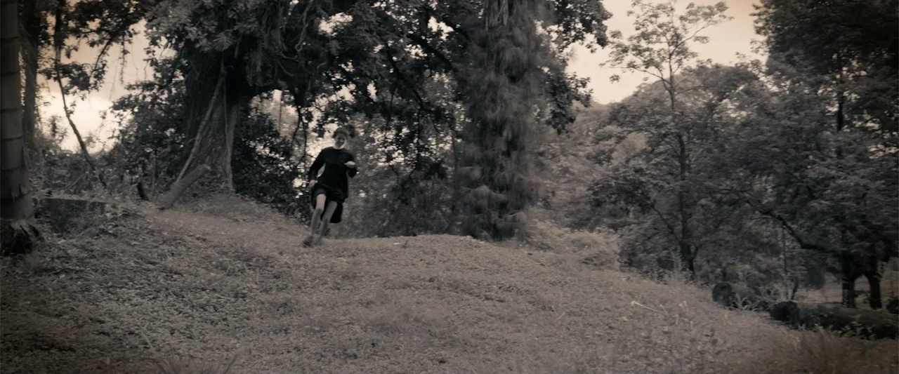 I Trust in You (2016) movie still: wide — A grassy, tree-filled hillside serves as the backdrop under a cloudy sky. A person in a dark dress moves…
