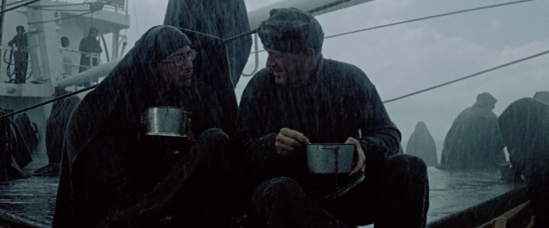 Papillon (1973) movie still: medium — A wet metal ship deck is populated by several men huddled in dark raincoats. Two men in the foreground sit and…