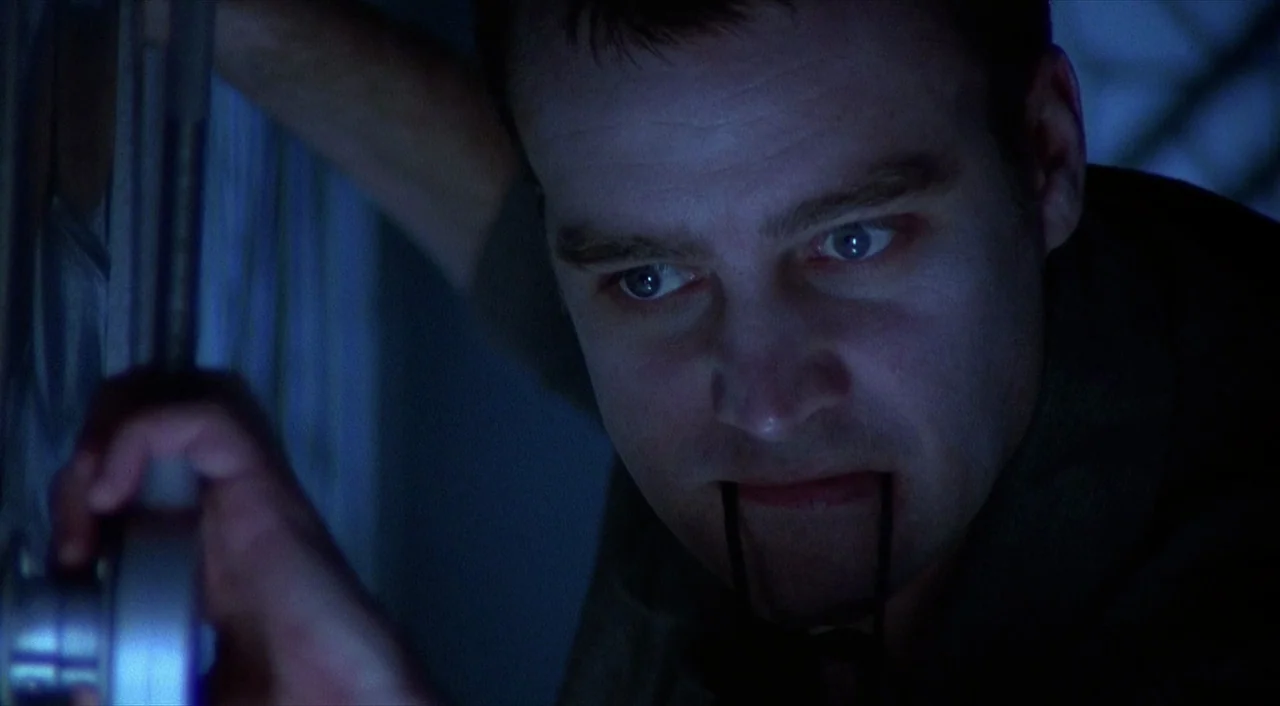 Cube (1997) movie still: closeup — A dark, dimly lit environment features a man peering through a doorway. He holds the bridge of his glasses…
