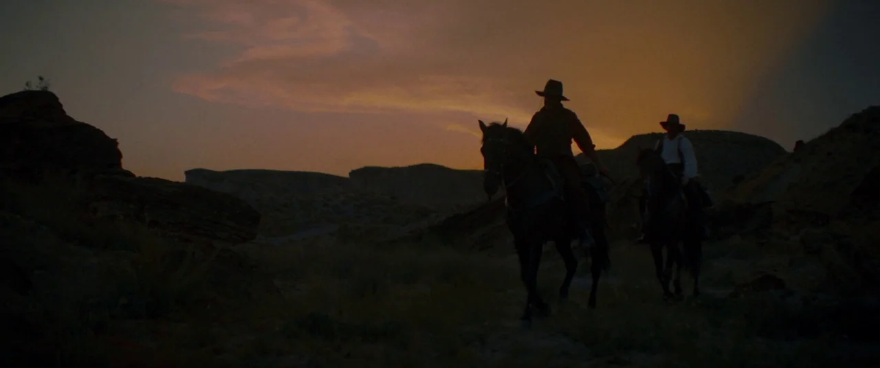 The Sisters Brothers (2018) movie still: wide — A desolate desert landscape with rocky outcrops at dusk under an orange sunset sky. Two men in…