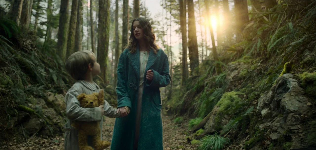 Peter Pan & Wendy (2023) movie still: fullbody — A dense forest path is lined with moss-covered rocks and tall evergreen trees. A young woman in a…