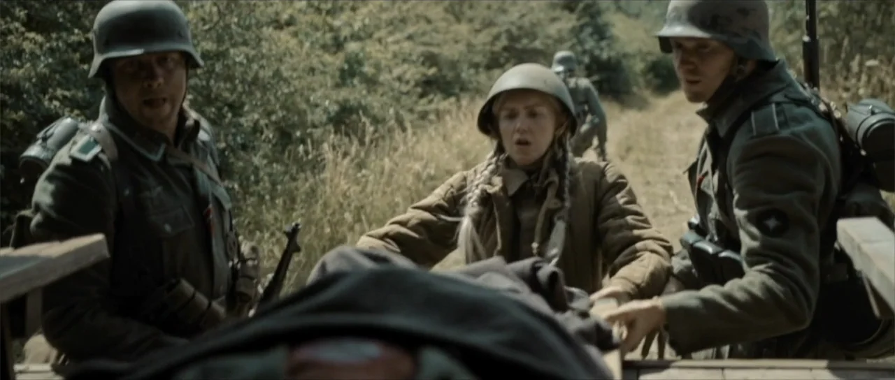 Sin retorno (2009) movie still: medium — A group of three soldiers carries a stretcher down a narrow, grass-lined dirt road. They wear 1940s-era…
