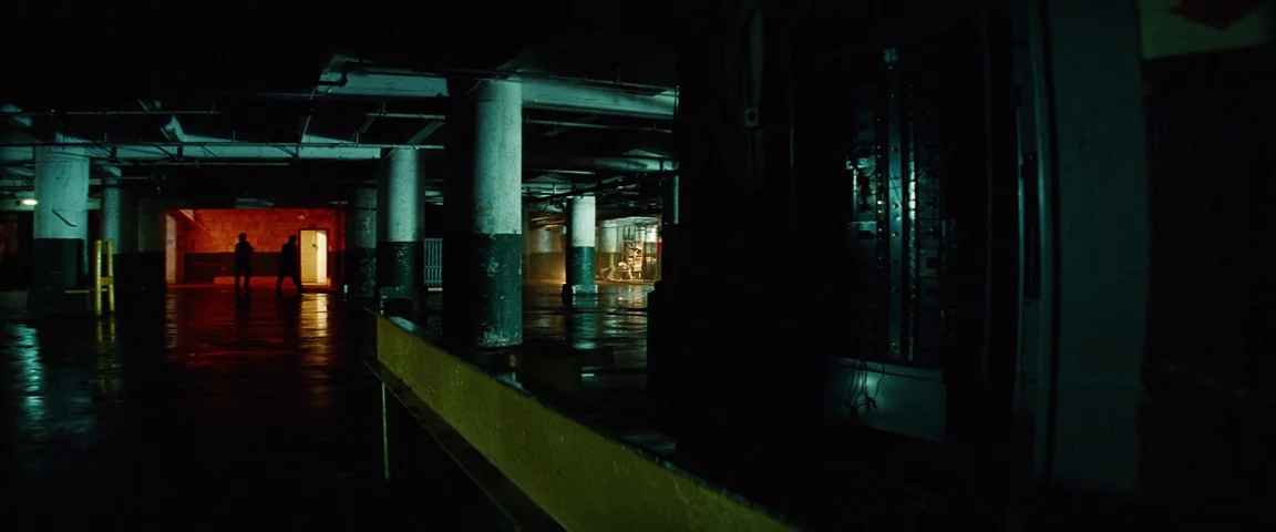 Savages (2012) movie still: wide — An expansive industrial parking structure features large concrete pillars and wet reflective floors. Two dark…