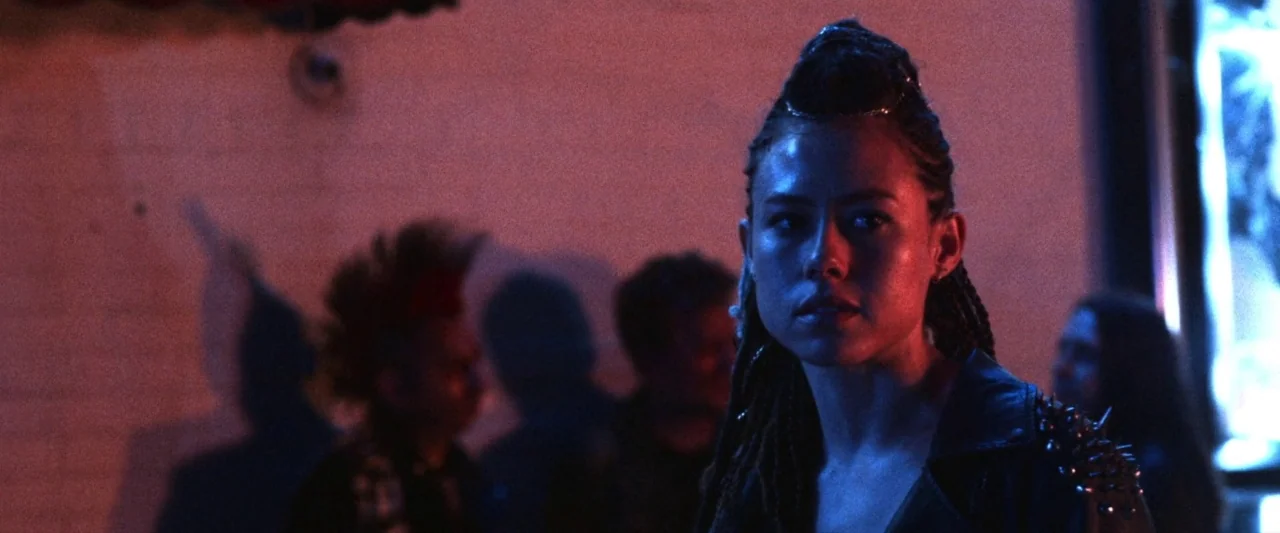 VFW movie still: medium — A dimly lit nightclub with brick walls illuminated by vibrant blue and red lighting. A young woman with braided hair…