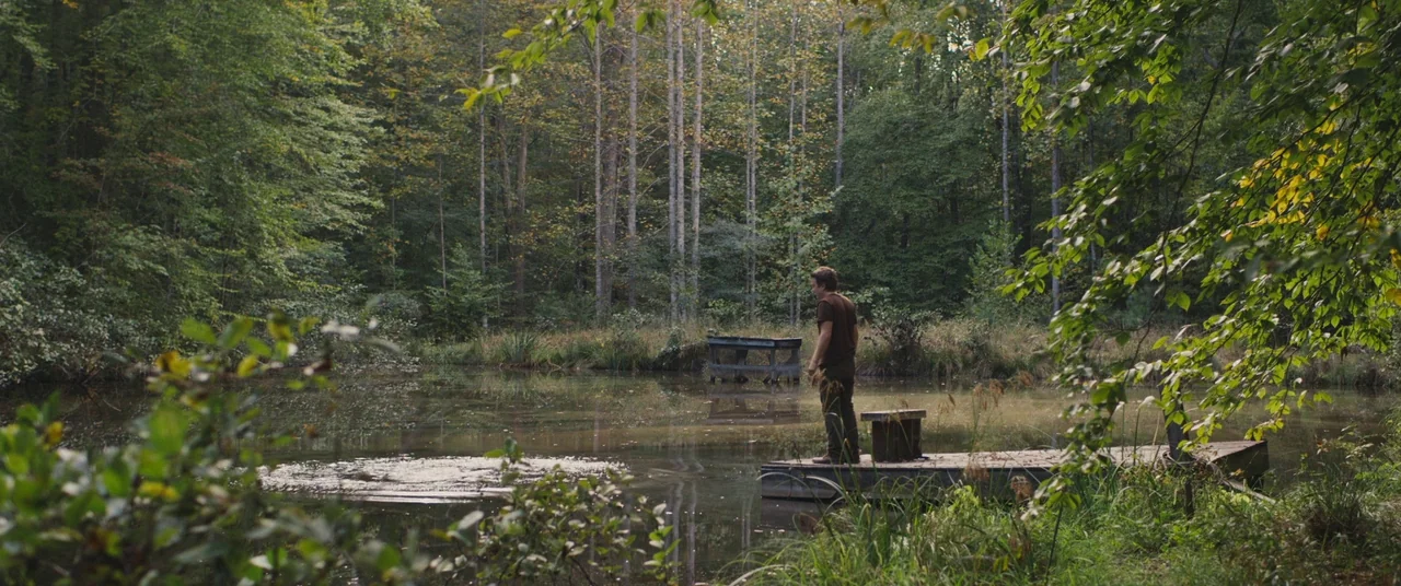 Blue Ruin (2013) movie still: wide — A dense forest borders a quiet pond with a wooden platform jutting out. A man stands on the platform looking…