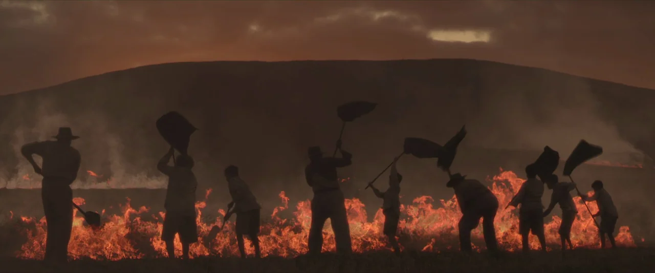The New Boy (2023) movie still: wide — A group of people works in a dark field during a grass fire. They use long tools to strike the flames and…
