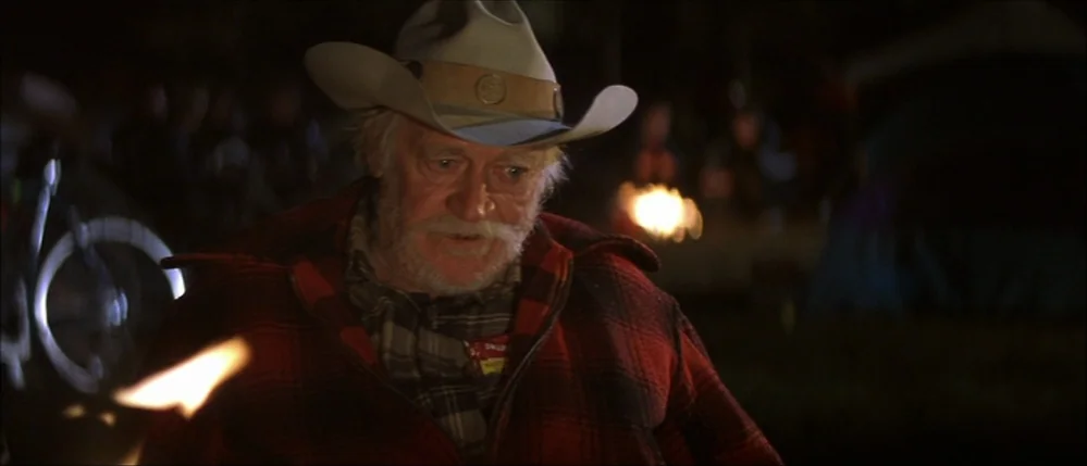 The Straight Story (1999) movie still: medium — An outdoor night scene at a campsite featuring a person near a fire. The elderly man speaks while…