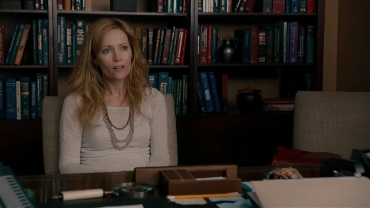 This Is 40 (2012) movie still: medium — An office with a wall of bookshelves filled with books behind a wooden desk. A woman sits in a chair at the…