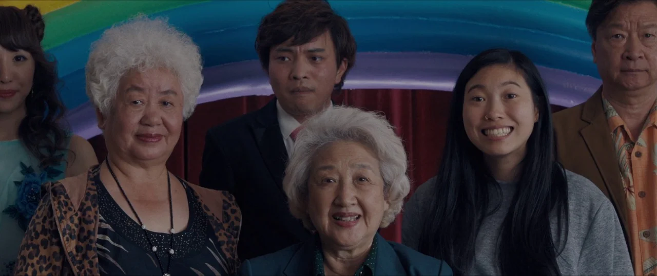 The Farewell (2019) movie still: medium — Five people stand positioned in a row against a decorative rainbow-colored backdrop. They pose together…