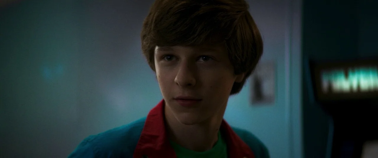 Summer of 84 (2018) movie still: closeup — A dimly lit indoor space features a teenager positioned near a glowing cabinet. The youth gazes towards…