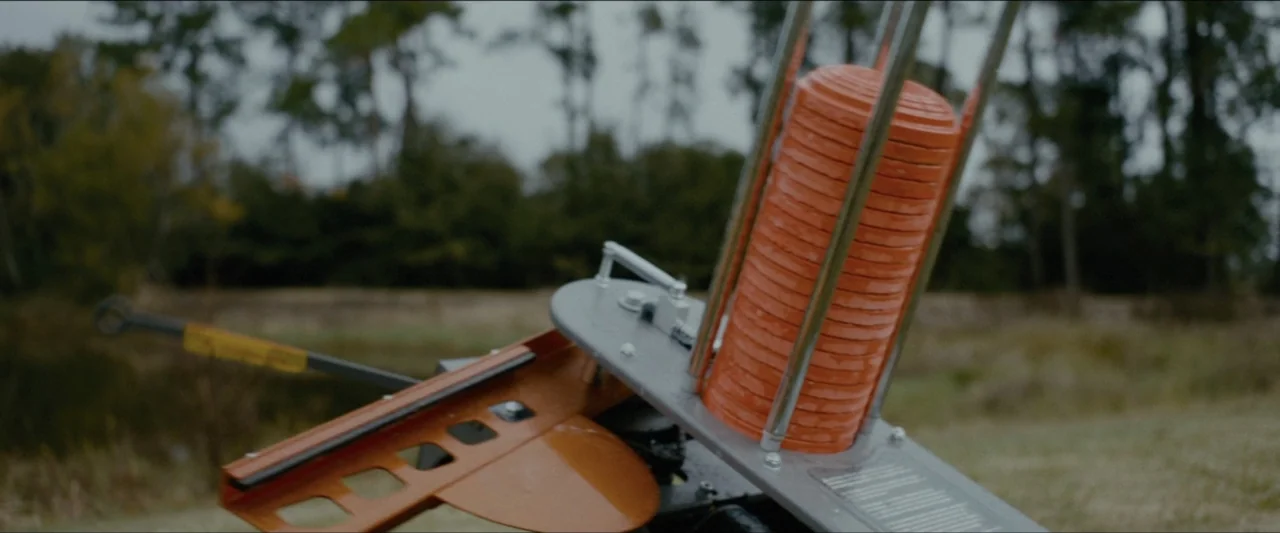 Ambush (2020) movie still: closeup — An outdoor field displays a mechanical clay target thrower set against a blurred treeline. A vertical stack of…