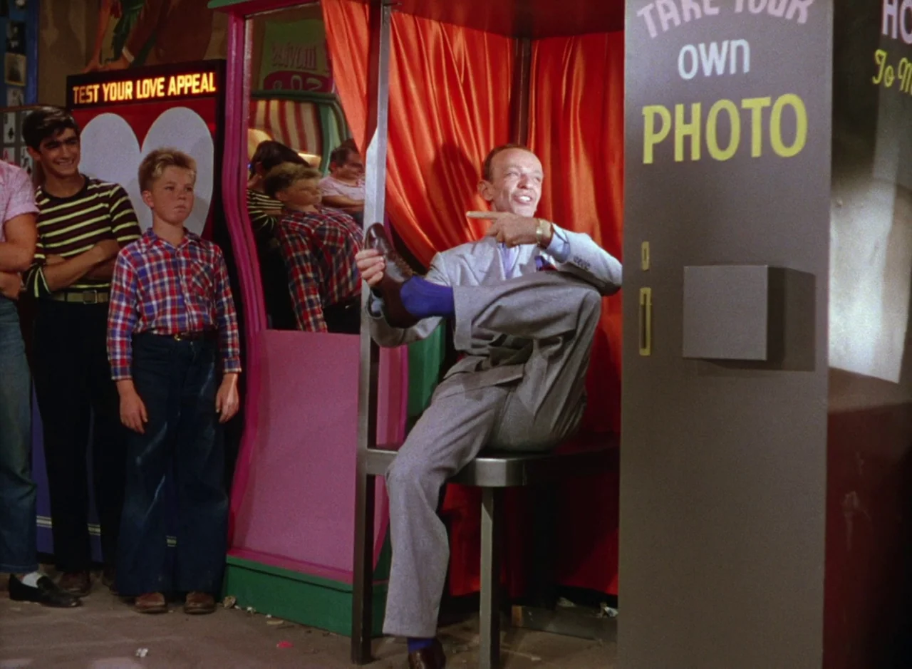 The Band Wagon (1953) movie still: medium — A brightly colored arcade interior featuring a photo booth and a love meter machine. An older man in a…