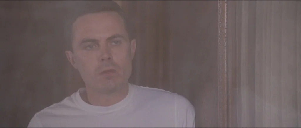 The Killer Inside Me (2010) movie still: closeup — A man stands in a dimly lit hallway with neutral-toned walls. The man is wearing a white t-shirt…