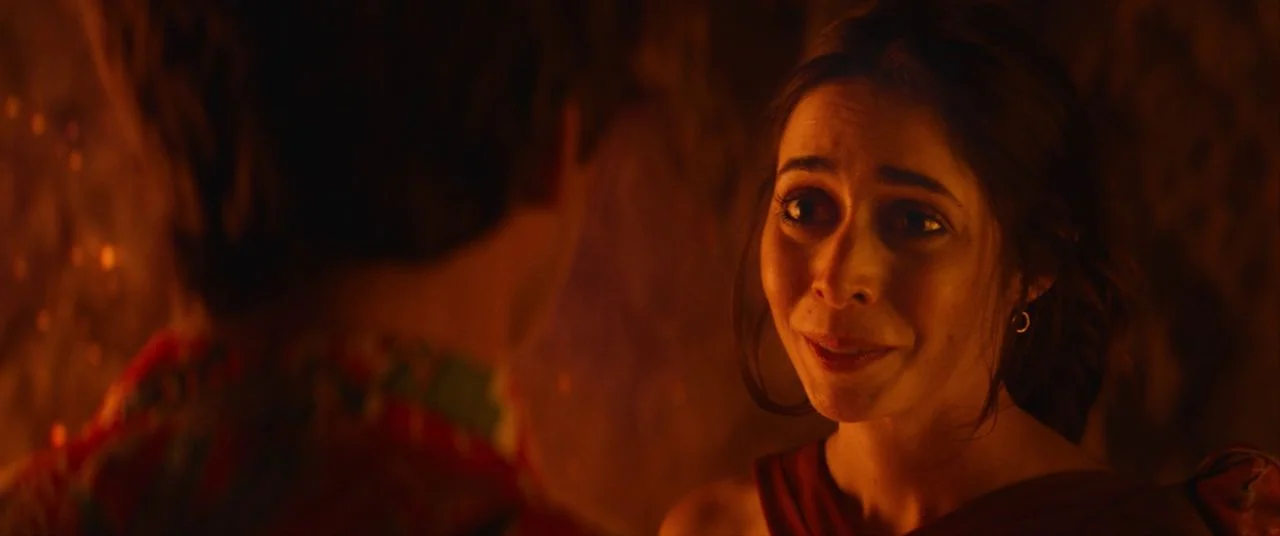 Palm Springs (2020) movie still: closeup — An interior space lit with warm tones suggests a social gathering environment. A woman with dark hair…