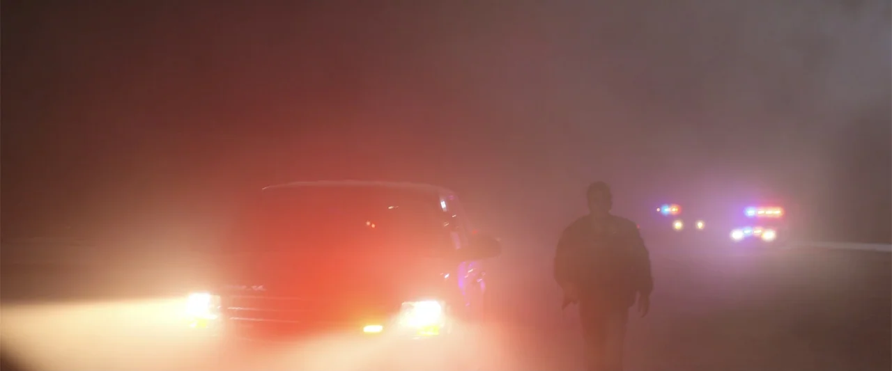 Trap House (2025) movie still: wide — A dark road is obscured by dense fog and illuminated by vehicle lights. A police officer walks toward the…