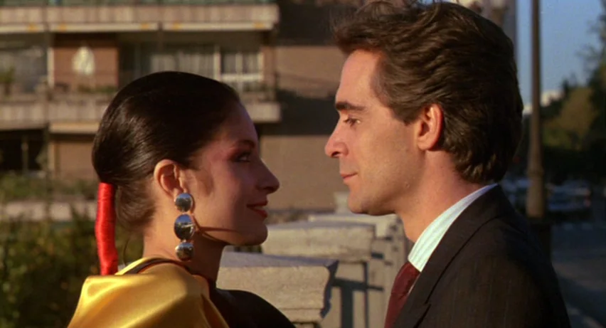 Matador (1986) movie still: medium — An urban sidewalk with a concrete balustrade and blurred apartment buildings in the background. A man in a suit…