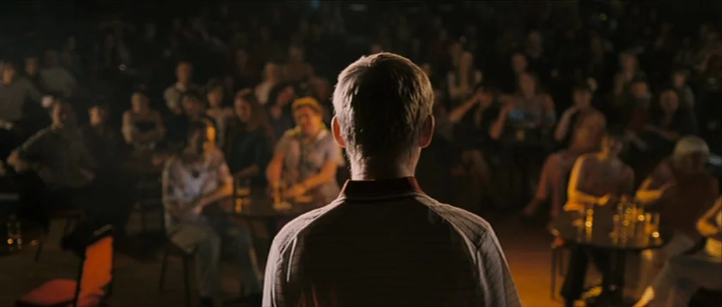 The Scouting Book for Boys (2009) movie still: medium — A dimly lit club audience sits at round tables in the shadowed background. A man wearing a…