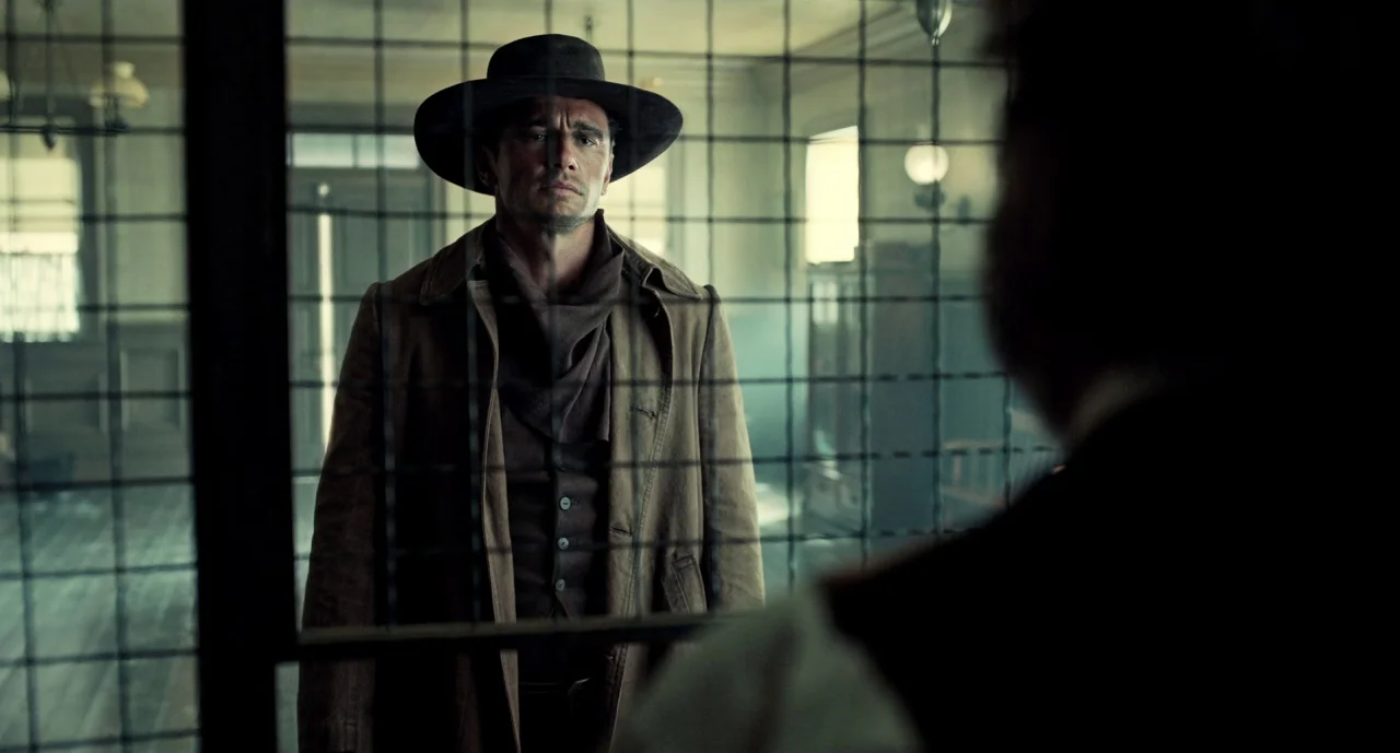 The Ballad of Buster Scruggs (2018) movie still: medium — A man in a cowboy hat stands behind a metal barred screen in an interior. The room behind…
