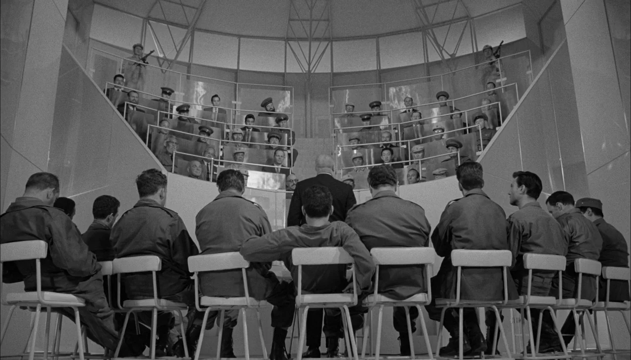 The Manchurian Candidate (1962) movie still: wide — A modernistic semi-circular room features tiered rows of men behind glass dividers. A group of…