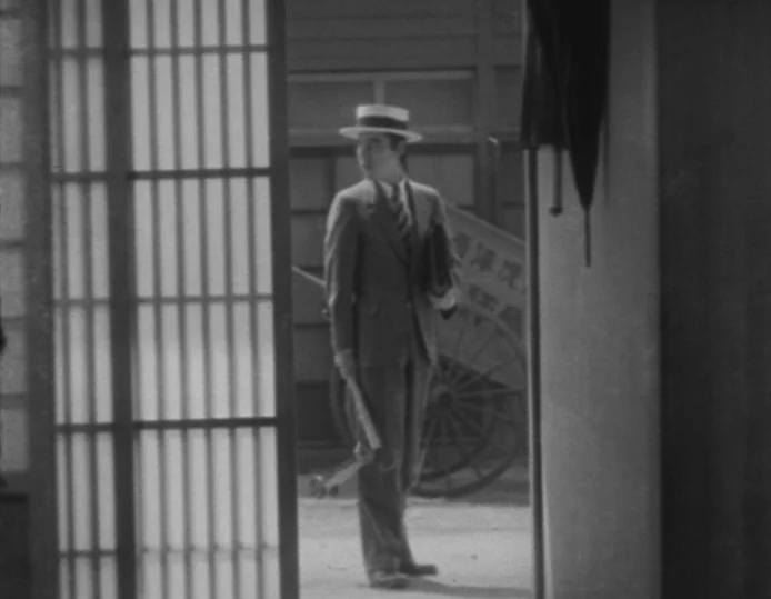 Tokyo Chorus (1931) movie still: fullbody — A man stands in the threshold of a Japanese building with paper sliding doors. Behind him is a…