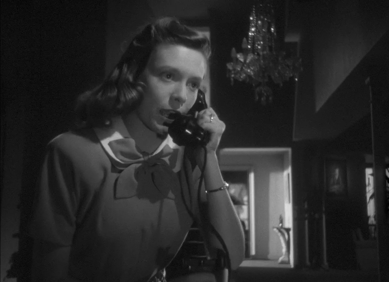 The Amazing Mr. X (1948) movie still: medium — A dimly lit room with a crystal chandelier hanging in the background. A woman is speaking into a…