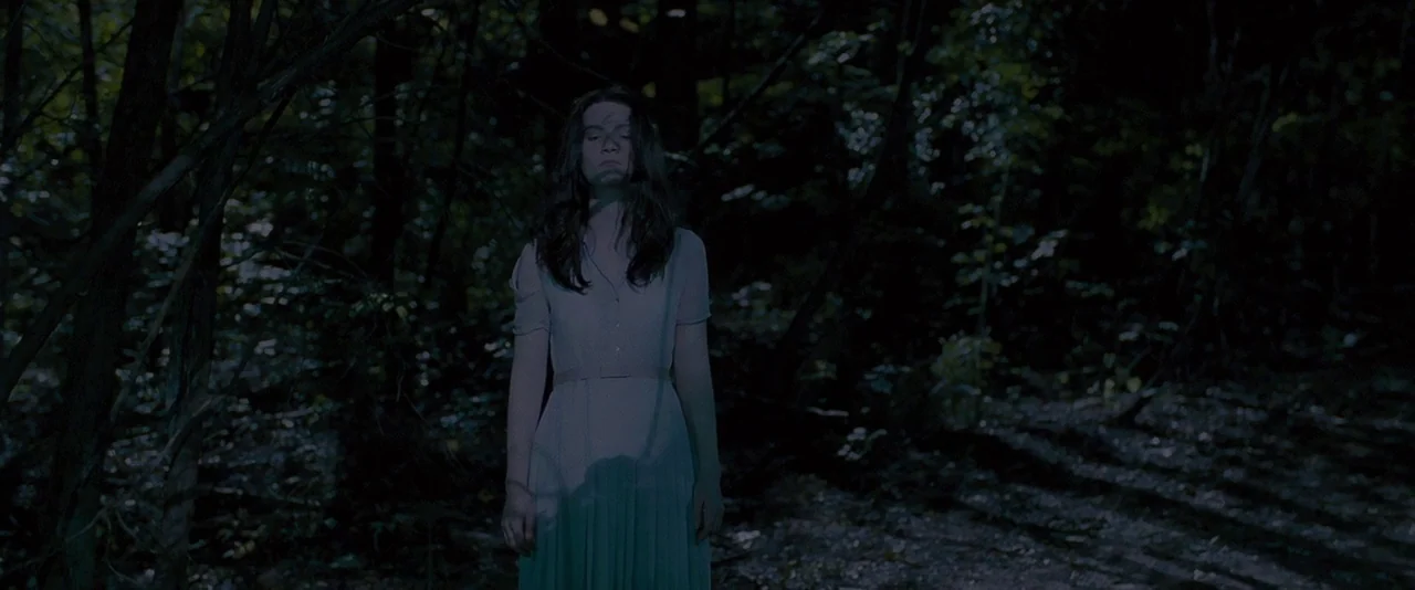 Stoker (2013) movie still: fullbody — A woman in a light-colored dress stands in a dark, tree-filled outdoor space at night. She remains motionless…