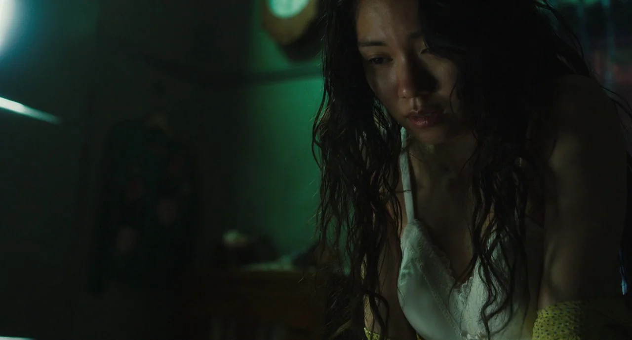 No Longer Human (2019) movie still: medium — A dimly lit room bathed in a distinct, murky green light at night. A young woman with long wavy hair…