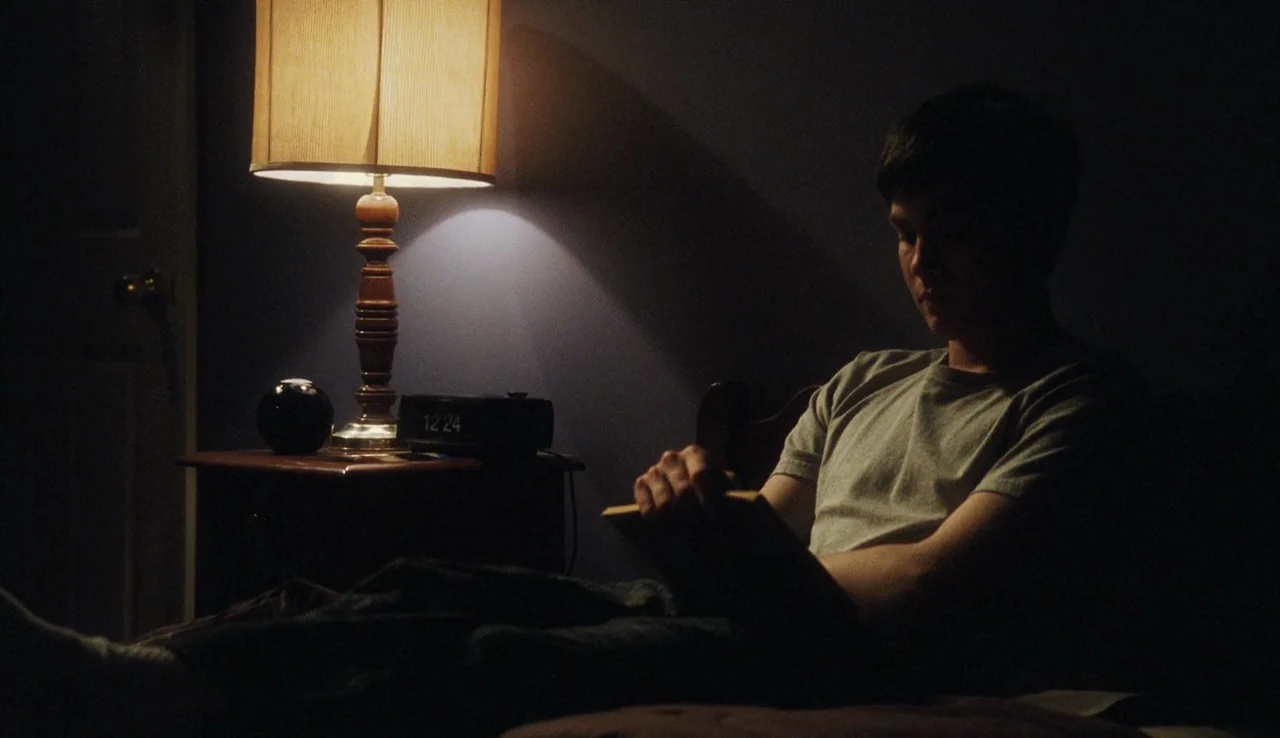 The Perks of Being A Wallflower (2012) movie still: medium — A dimly lit room with dark walls features a nightstand holding a lit lamp. A young man…