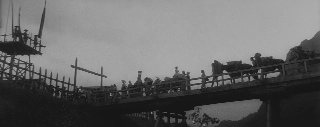 The Hidden Fortress (1958) movie still: wide — A group of people and loaded pack animals cross a weathered wooden bridge. The procession travels…