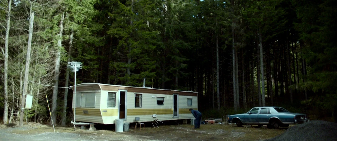 The House That Jack Built (2018) movie still: wide — A vintage cream and brown mobile home is situated in a dense coniferous forest. A lone man…