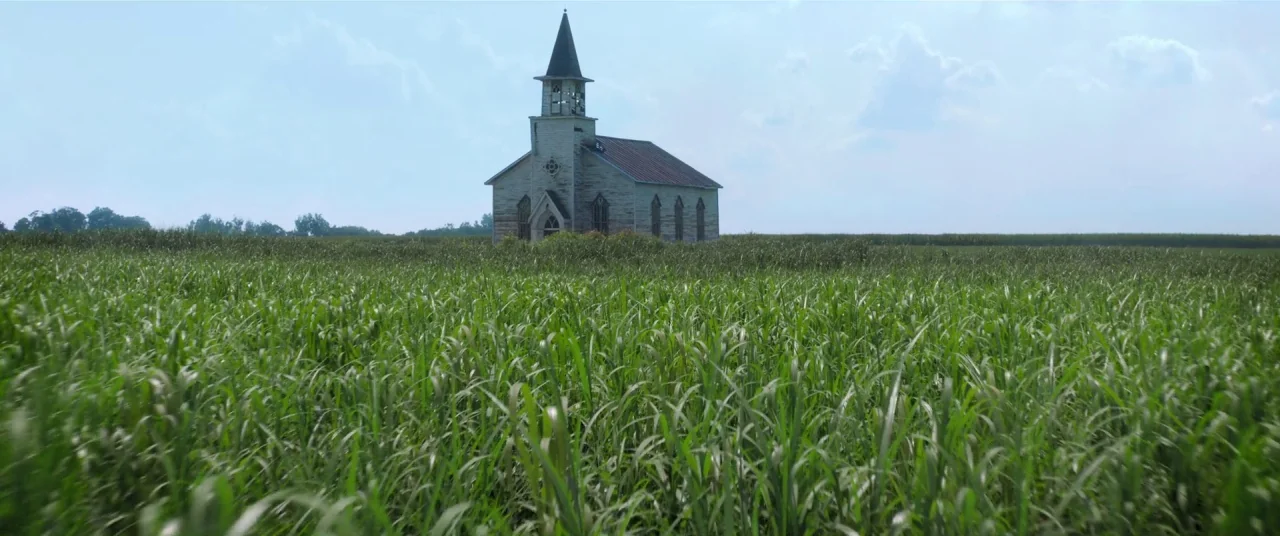 In The Tall Grass (2019) movie still: establishing — A small weathered white church with a spire sits amidst a large green field. Dense tall grass…