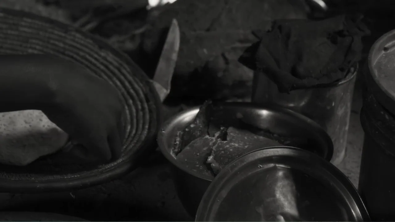 Faya Dayi (2021) movie still: closeup — A dark indoor space contains a metal cooking bowl filled with food pieces. A single hand reaches into a…