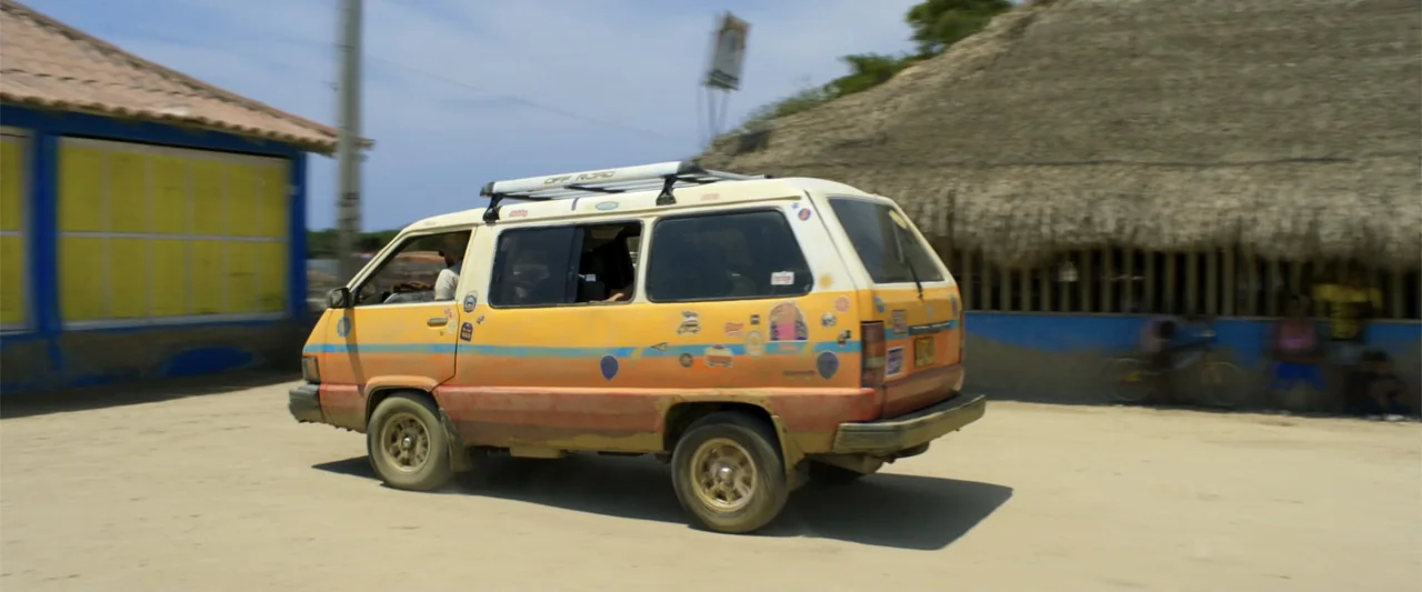 Férias Trocadas (2024) movie still: medium — A yellow and orange van travels along a sandy, sunlit road near a thatched building. The vehicle…