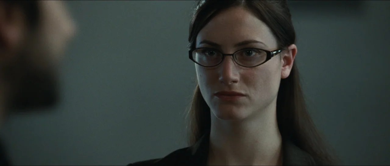 Slackers (2002) movie still: closeup — An interior office space with neutral, grey-toned walls and professional furniture. A woman with glasses…