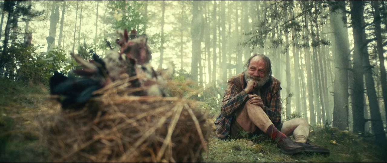The Toxic Avenger (2023) movie still: medium — A dense coniferous forest features thick hazy air and tall slender tree trunks. An elderly man sits…