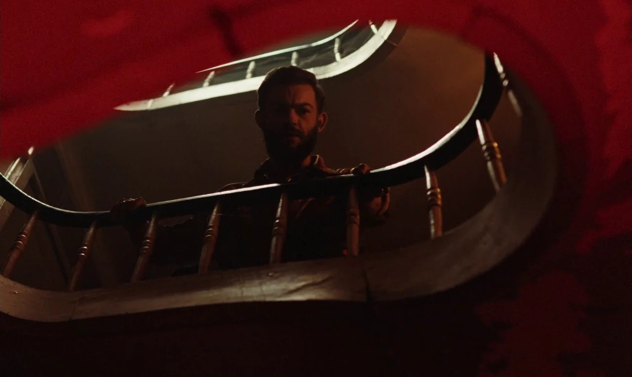 Maldoror (2024) movie still: medium — A man stands behind a curved wooden bannister in a dimly lit, red-saturated space. He looks down from the…