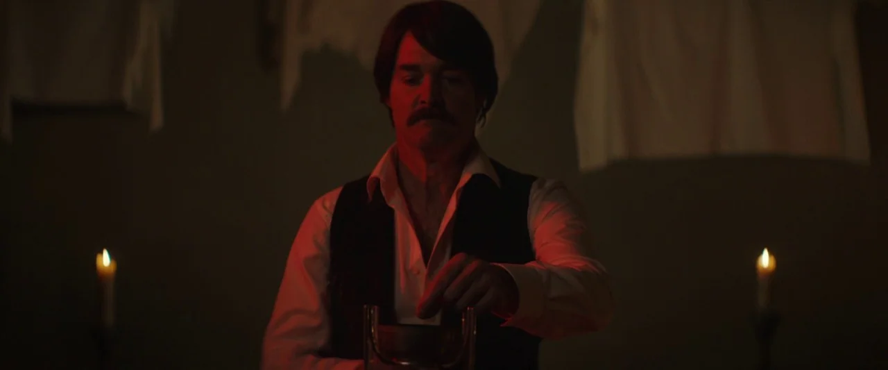 Extra Ordinary (2019) movie still: medium — A dimly lit room features white cloths hanging on the dark wall behind a man. The man sits at a table…