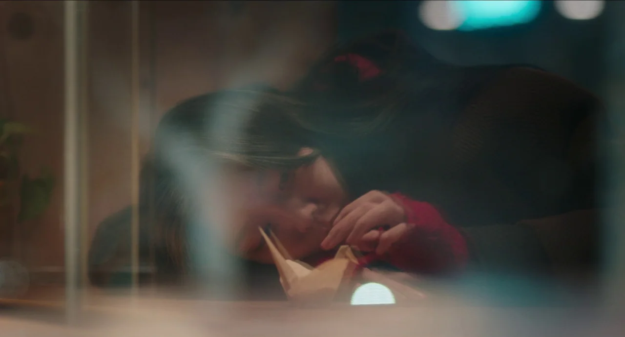Microhabitat (2017) movie still: closeup — A person rests their head on a surface next to a paper origami crane. The view is partially obscured by a…