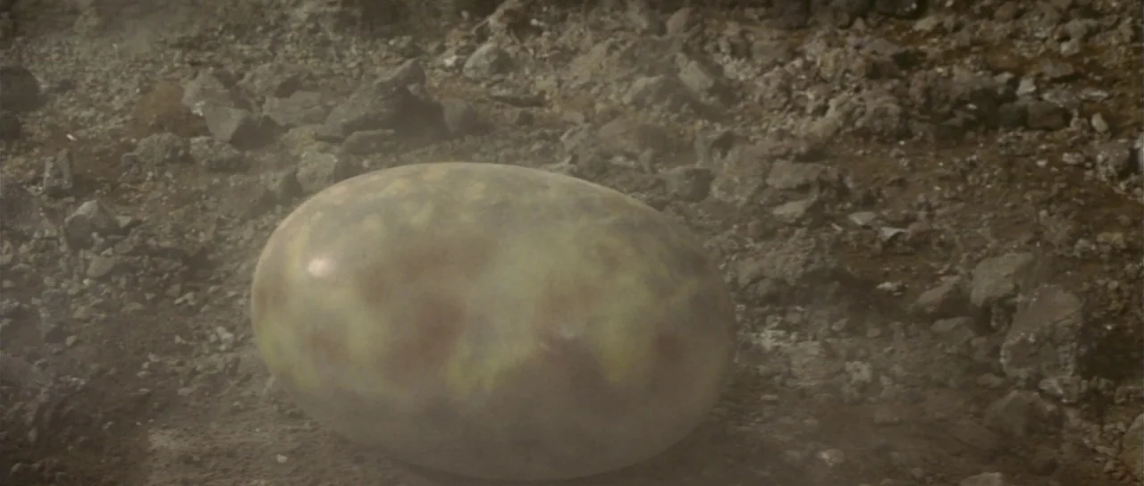 Son of Godzilla (1967) movie still: closeup — A smooth, oblong, semi-translucent egg rests in a patch of loose dirt and rocks. Thin dust or mist…