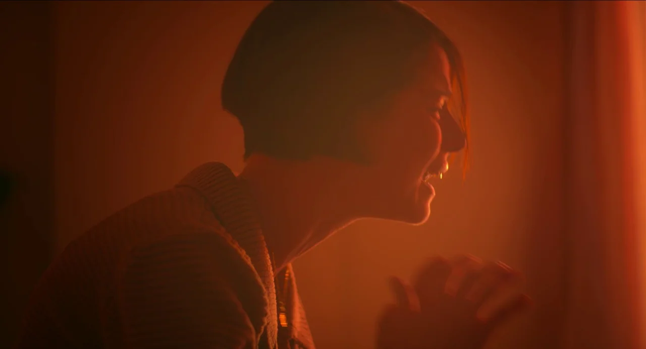 Men (2022) movie still: medium — A dimly lit indoor space glows with a saturated amber light. A young woman with a short haircut shouts with her…