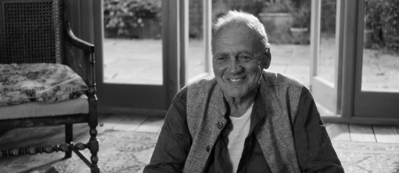 The Party (2017) movie still: medium — An elderly man sits in a sunlit room featuring large glass doors. He looks forward with a smile while wearing…