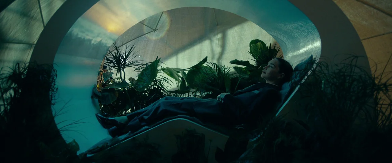 The Pod Generation (2023) movie still: medium — A person rests inside a circular architectural pod filled with lush green indoor tropical plants.…