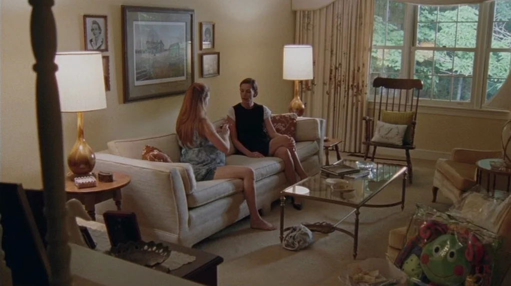 Junebug (2005) movie still: wide — A well-furnished room contains a beige sofa, glass coffee table, and table lamps. Two women are sitting together…