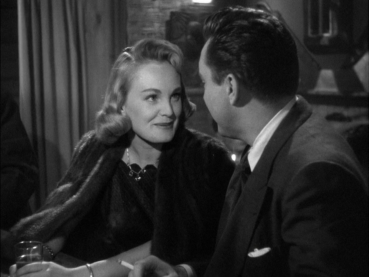 D.O.A. (1949) movie still: medium — A dimly lit club with dark curtains and textured wall elements in the background. A woman sits across from a…