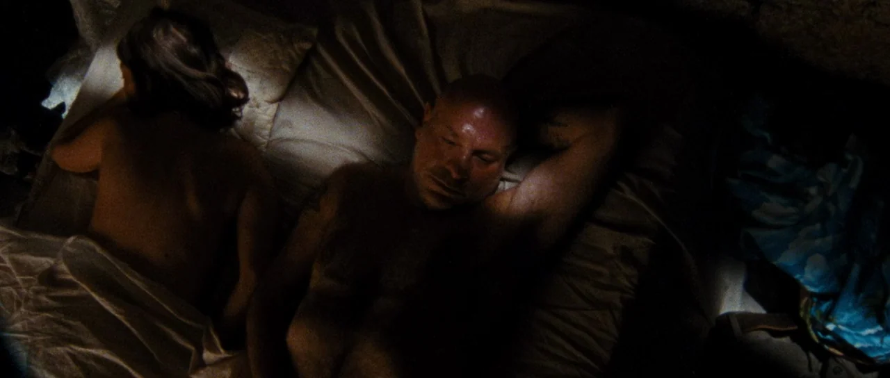 Let The Corpses Tan (2017) movie still: medium — A dark bedroom with white linens covering a couple on a mattress. The man and woman are lying…