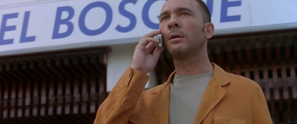 Talk To Her (2002) movie still: medium — A man stands outdoors in front of a white sign with blue text. He holds a silver mobile phone to his ear.