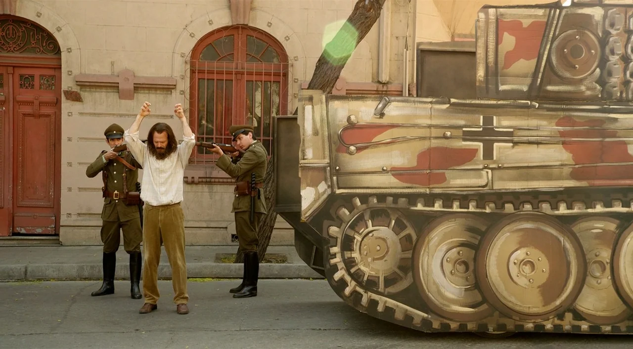 Dance of Reality (2013) movie still: wide — A man stands on a city street corner with his hands held up high. Two uniformed soldiers hold rifles…
