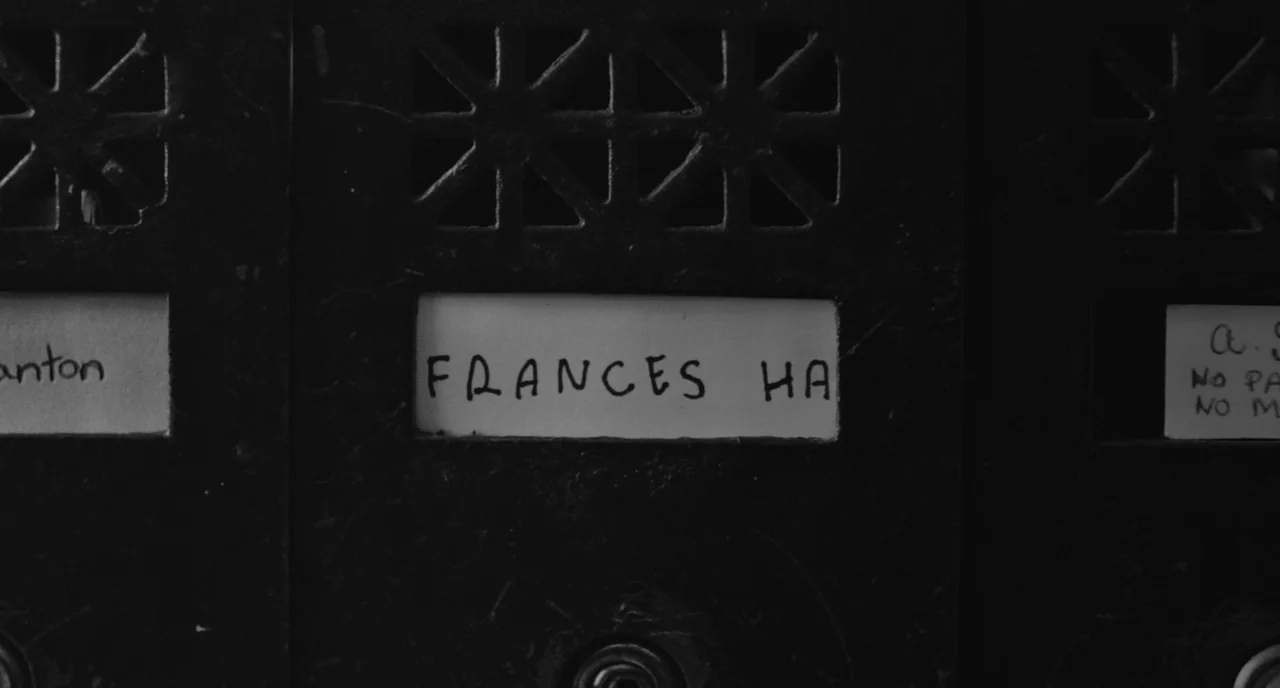 Frances Ha (2012) movie still: closeup — A set of black metal mailboxes with slotted vent patterns sits in an interior. A white paper label with…