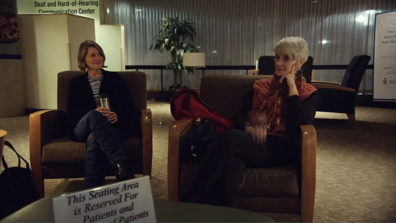 Suited (2016) movie still: medium — A quiet waiting area features carpeted floors and beige chairs in a dimly lit space. Two women sit in individual…