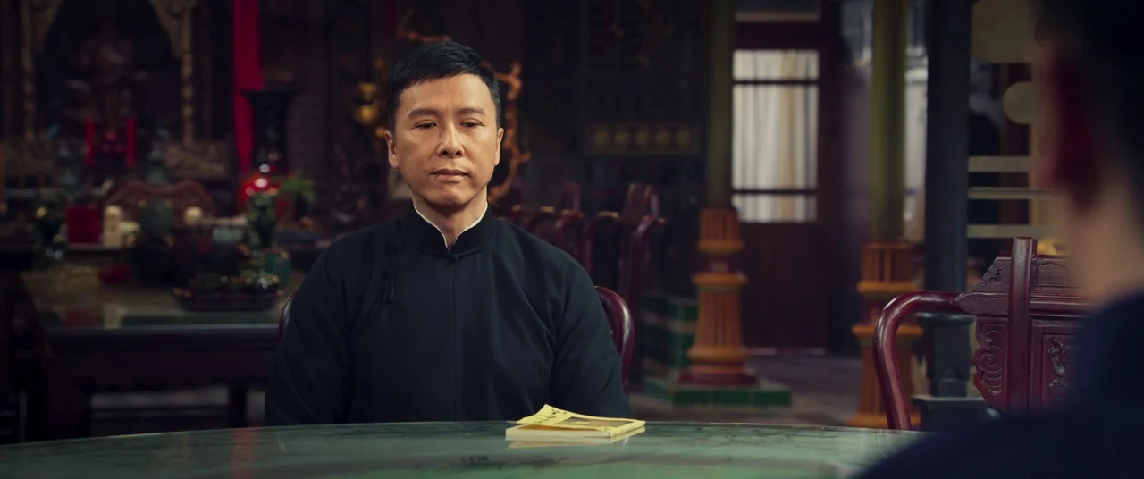 Ip Man 4: The Finale (2019) movie still: medium — A traditional room with carved wooden furniture and a polished round stone-top table. A man in a…